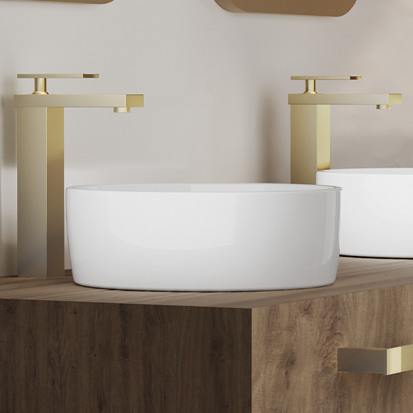 Grifería de baño lujoso color dorado de la colección Limited de Helvex, acompañada del lavabo morea circular color blanco encima de un vanity de madera